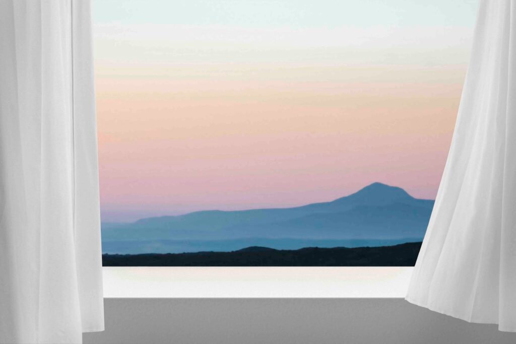 View through white curtains revealing layered mountains and a soft pastel sunrise in the distance
